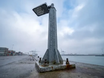 Stalen lichtmast met geboute voetplaat op industrieel betonnen oppervlak in Nederlandse havenzone, gefotografeerd vanuit lage hoek.
