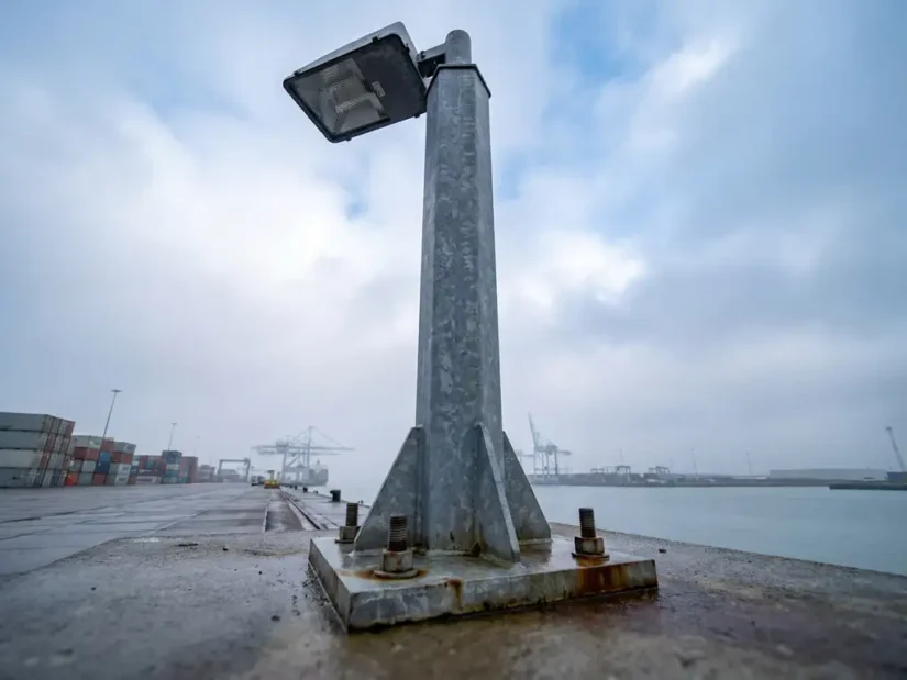 Stalen lichtmast met geboute voetplaat op industrieel betonnen oppervlak in Nederlandse havenzone, gefotografeerd vanuit lage hoek.