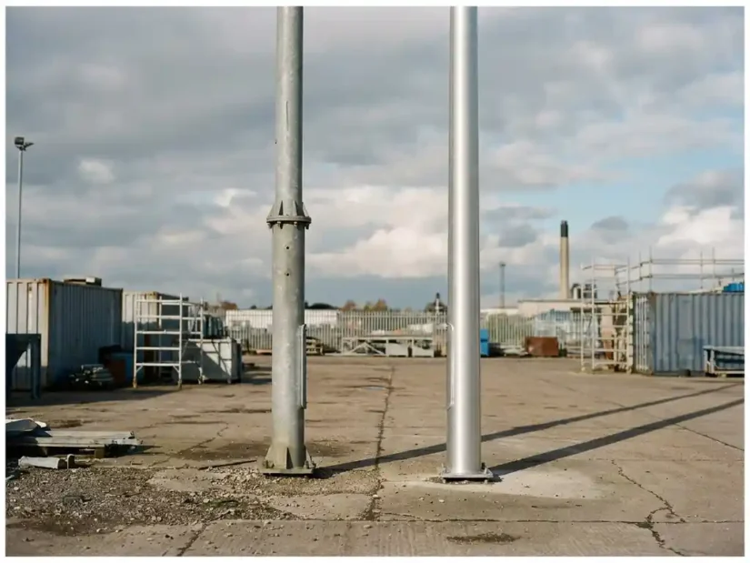 Twee industriële lichtmasten naast elkaar, één met mat grijs staal en één met geborsteld aluminium, in natuurlijk daglicht op betonnen ondergrond.