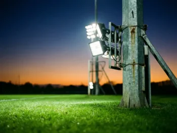Verlichte stalen lichtmast op een sportveld bij schemering, met felle witte floodlights boven groen gras en een diepblauwe lucht.