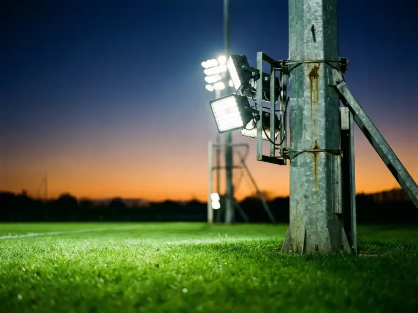 Verlichte stalen lichtmast op een sportveld bij schemering, met felle witte floodlights boven groen gras en een diepblauwe lucht.