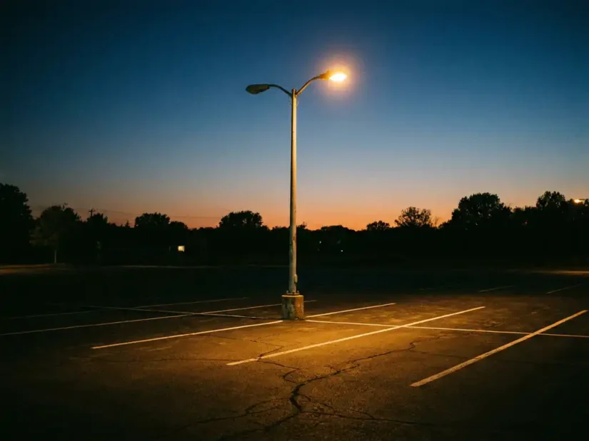 Stalen lichtmast in leeg parkeerterrein bij schemering, met warm amberkleurig straatlicht tegen een blauwe avondlucht.