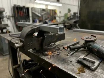 Unistrut kanaalprofiel vastgeklemd in bankschroef, omgeven door staalvijlsel en vonken op een industriële werkbank.